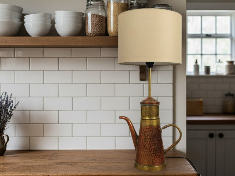 A unique, handmade vintage copper and brass coffee pot repurposed into a table lamp with a cream shade, placed on a wood kitchen counter with white subway tile backsplash. Perfect as a cool desk lamp or accent light.