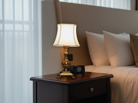 Vintage ornate brass table lamp with off-white shade on a nightstand. Perfect mid-century bedside or accent light.