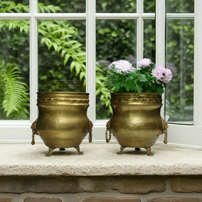 A pair of vintage hammered brass jardiniere vessels featuring intricate lion head ring handles, a decorative rope-twist rim, and raised tripod claw feet. 